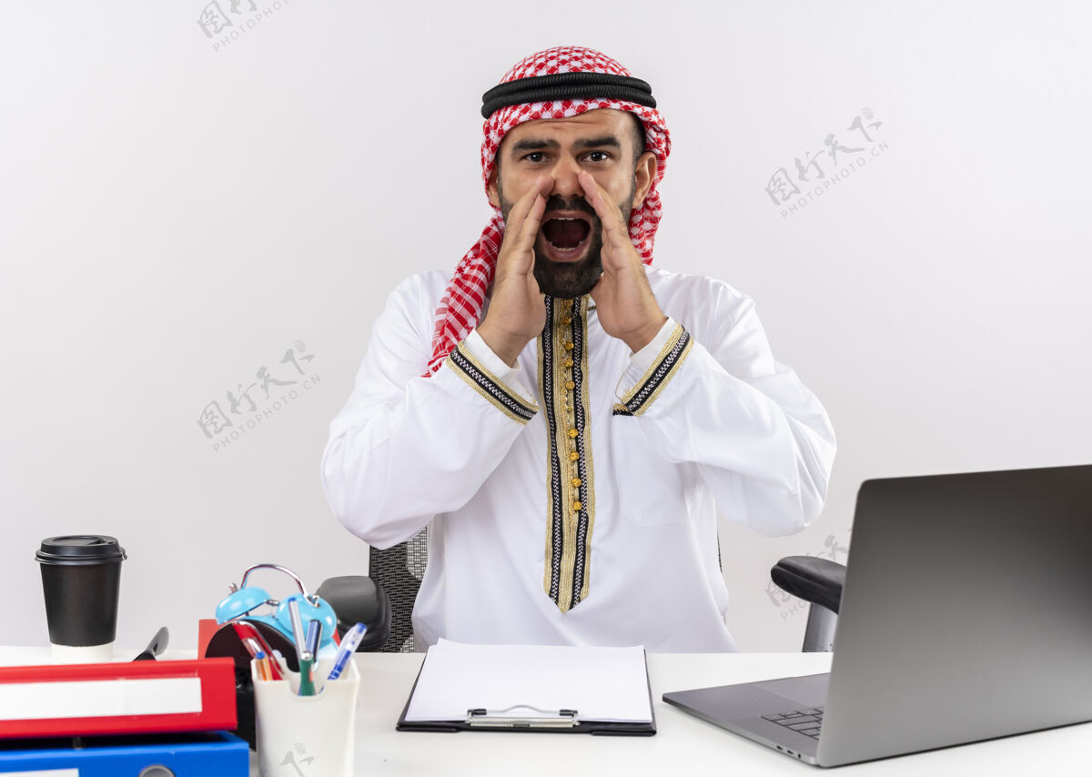 笔记本电脑身着传统服装的阿拉伯商人坐在桌旁用笔记本电脑大声叫喊 用手靠近嘴巴在办公室工作坐电脑手