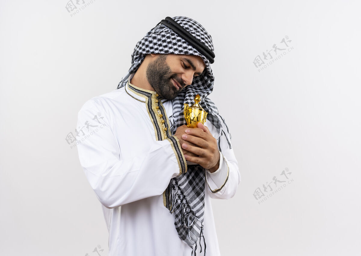 阿拉伯语身着传统服装的阿拉伯男子站在白墙上拥抱奖杯 微笑着感受积极的情绪传统拥抱男人