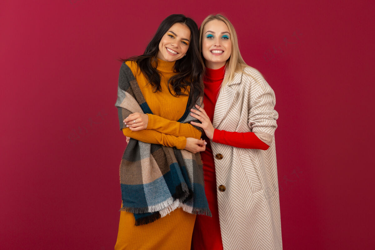 冷两位笑容可掬的时尚女性穿着秋冬时装和外套 在红墙上孤零零地摆着姿势秋天女人女士