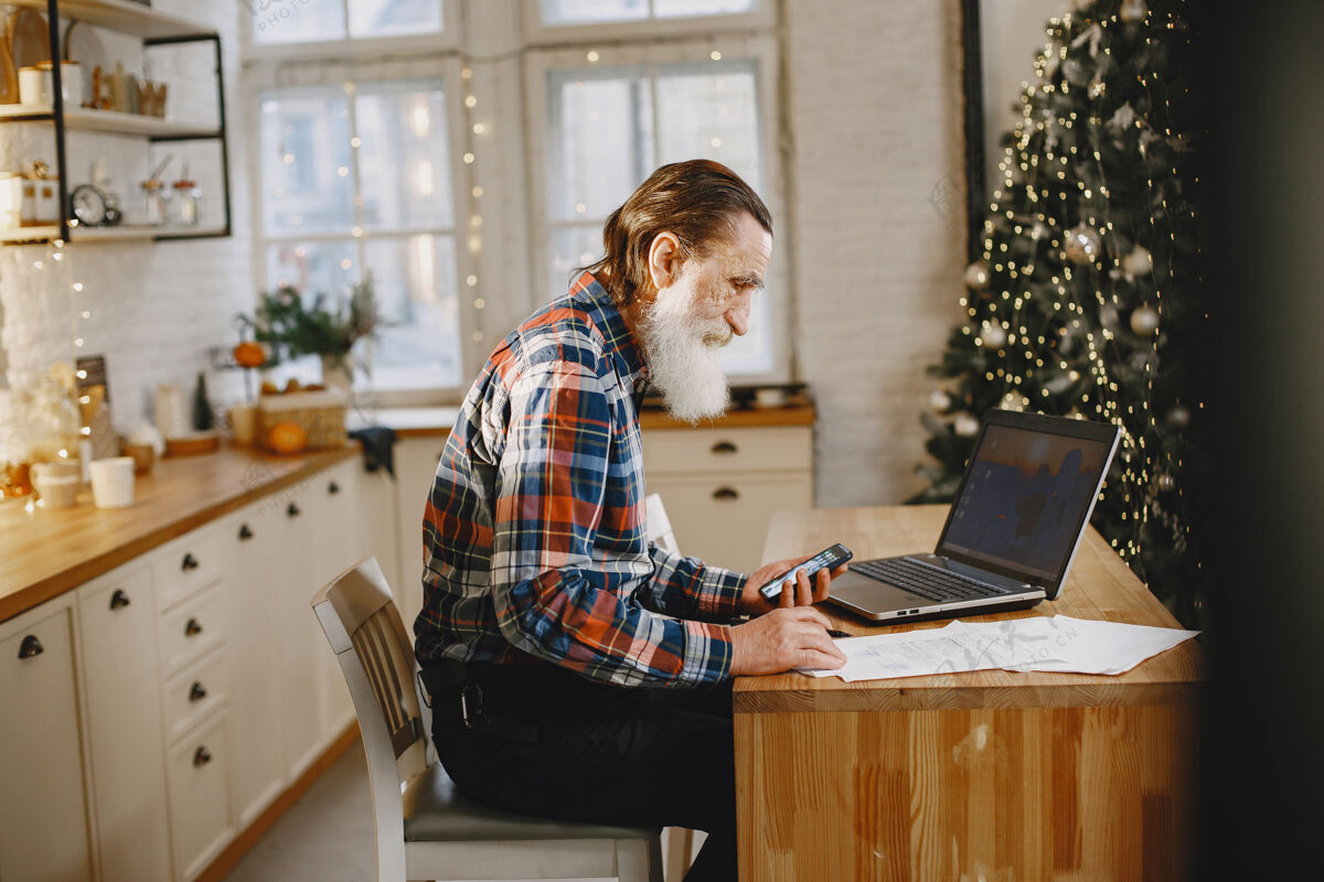 胡须老人拿着笔记本电脑爷爷坐在圣诞装饰品里老人拿着手机房子笔记本电脑家庭