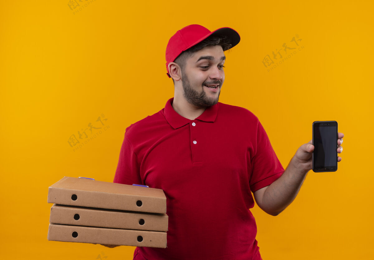 披萨年轻的送货员穿着红色制服 戴着帽子 手里拿着一叠比萨饼盒 脸上带着微笑看着它制服帽子红色