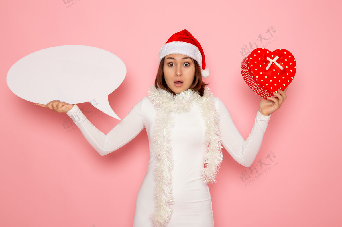 颜色正面图年轻女子手持白色大招牌 在粉色墙面上呈现彩雪圣诞新年情感假期圣诞节情感大