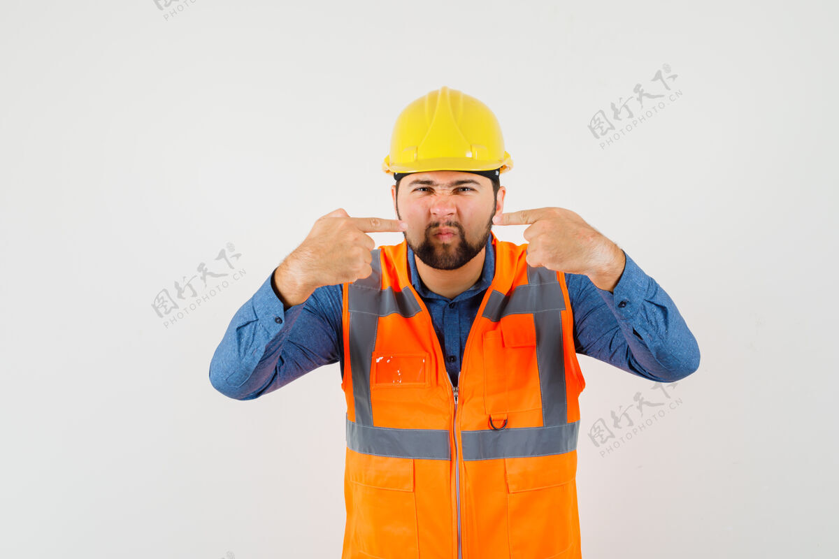 工厂年轻的建筑工人穿着衬衫 背心 头盔指着他的鼻子 同时皱眉 前视图工厂安全帽车间