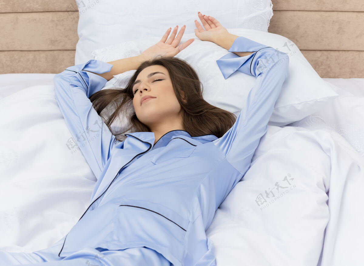 睡衣穿着蓝色睡衣的年轻漂亮女人躺在床上 躺在柔软的枕头上 安详地睡在家里卧室的室内灯光背景下和平房子卧室