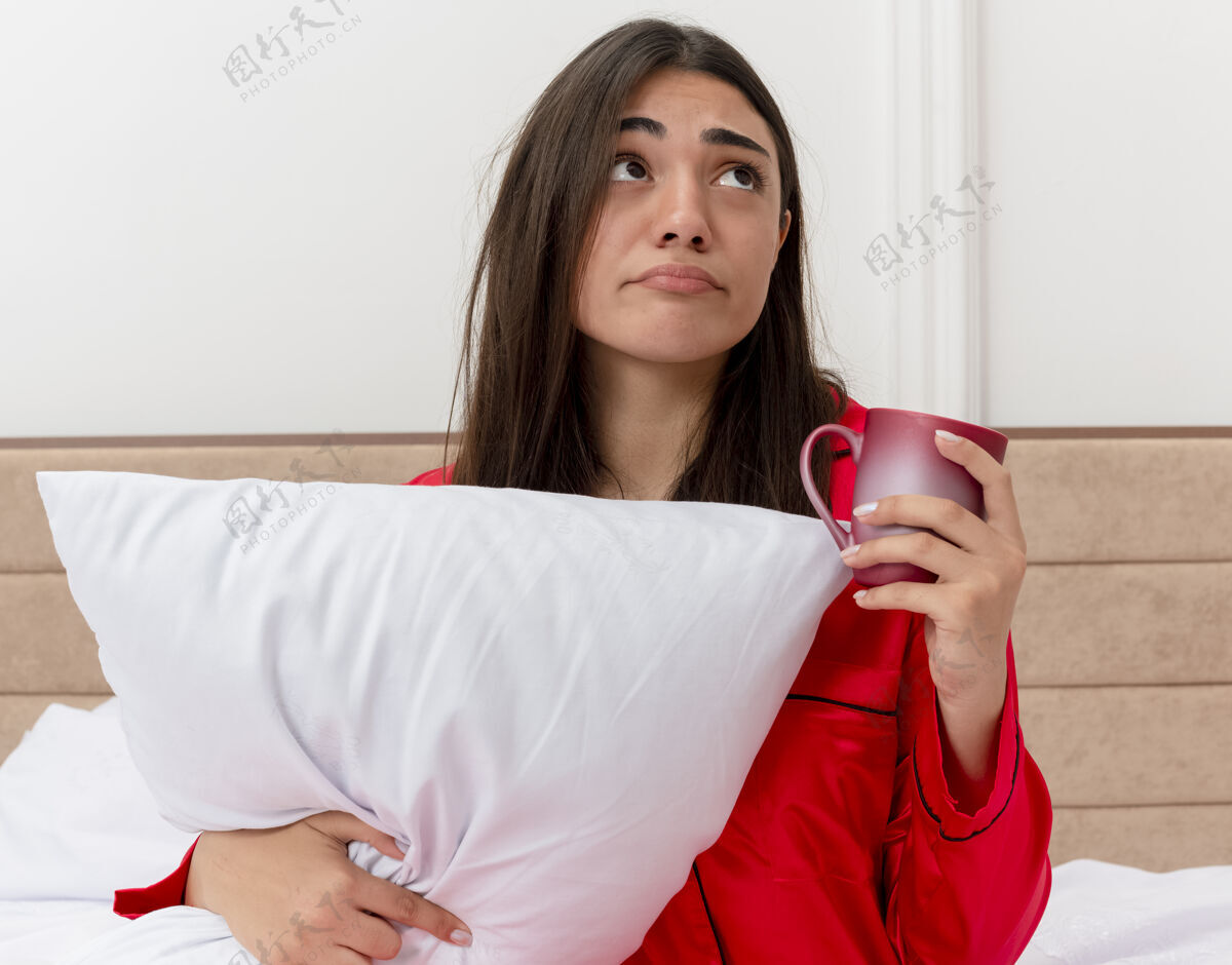 女人穿着红色睡衣的年轻美女坐在床上 枕着一个杯子 抬头望着卧室里的灯光背景 感到不快和困惑起来看着杯子