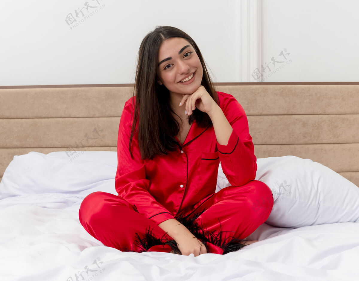 快乐穿着红色睡衣的年轻漂亮女人坐在床上看着摄像机 在灯光的衬托下 她在卧室的内部笑容可掬坐着睡衣女人