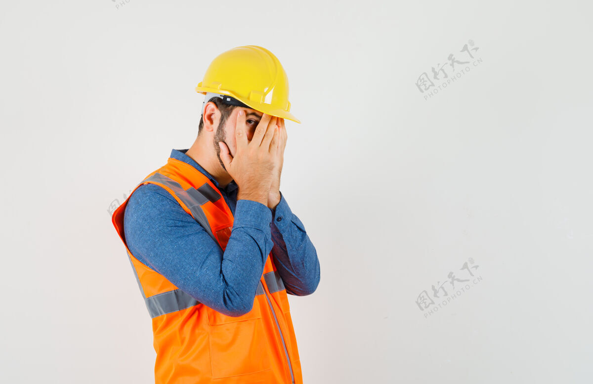 安全年轻的建筑工人穿着衬衫 背心 头盔透过手指看 看起来很害怕 前视图制服专业工具