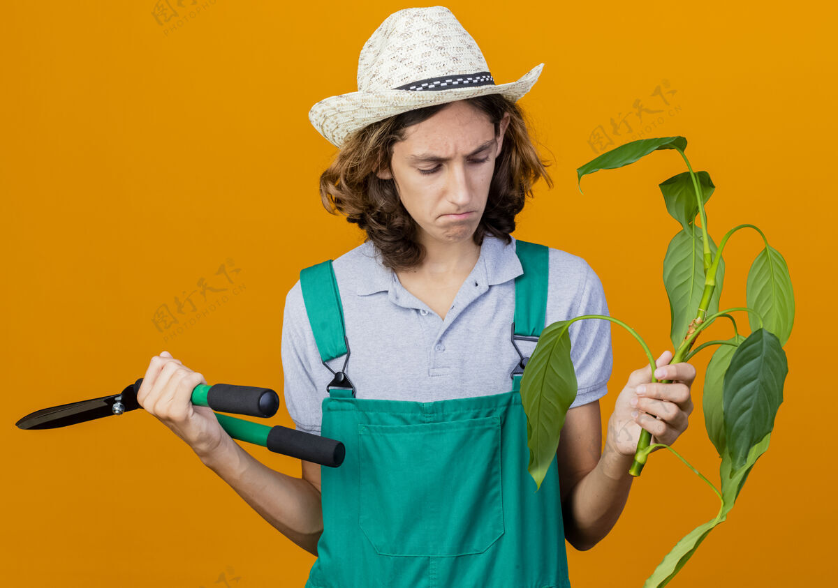 男人年轻的园丁穿着连体衣戴着帽子拿着植物和树篱剪帽子篱笆悲伤