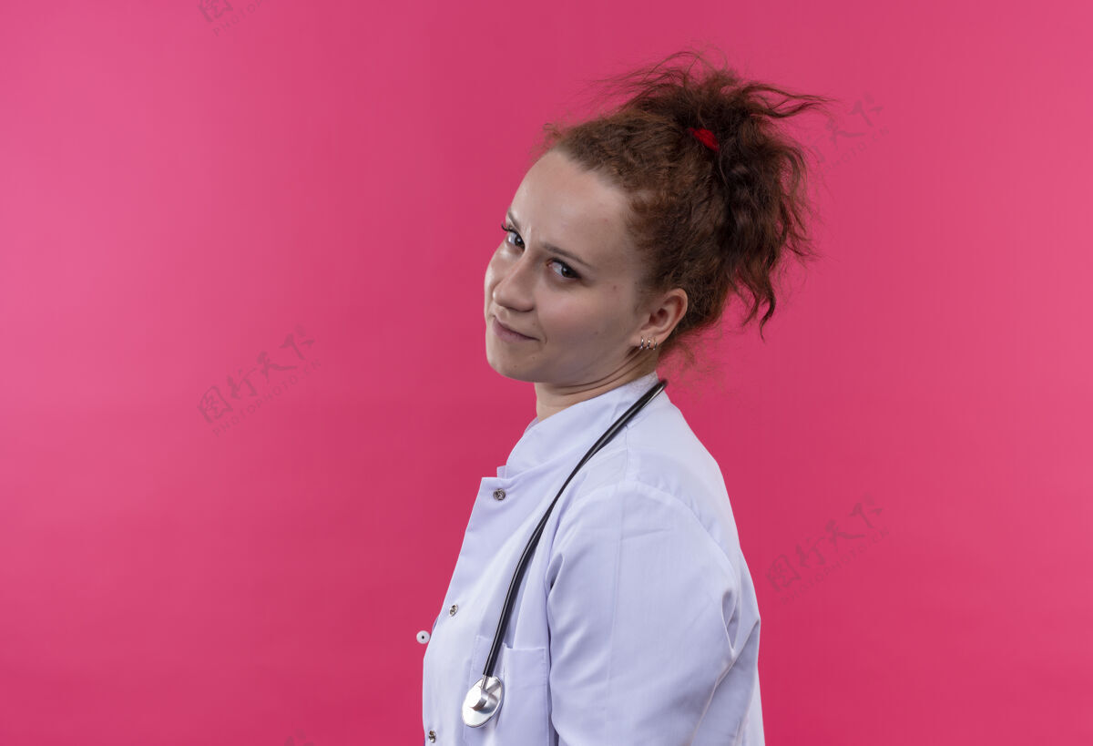外套年轻的女医生穿着白大褂 带着听诊器 带着怀疑的表情站在粉色的墙上侧着怀疑表情