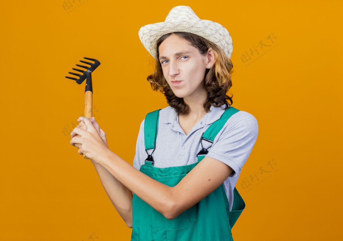微笑年轻的园丁 穿着连体衣 戴着帽子 摇着小耙子男人连身衣耙子