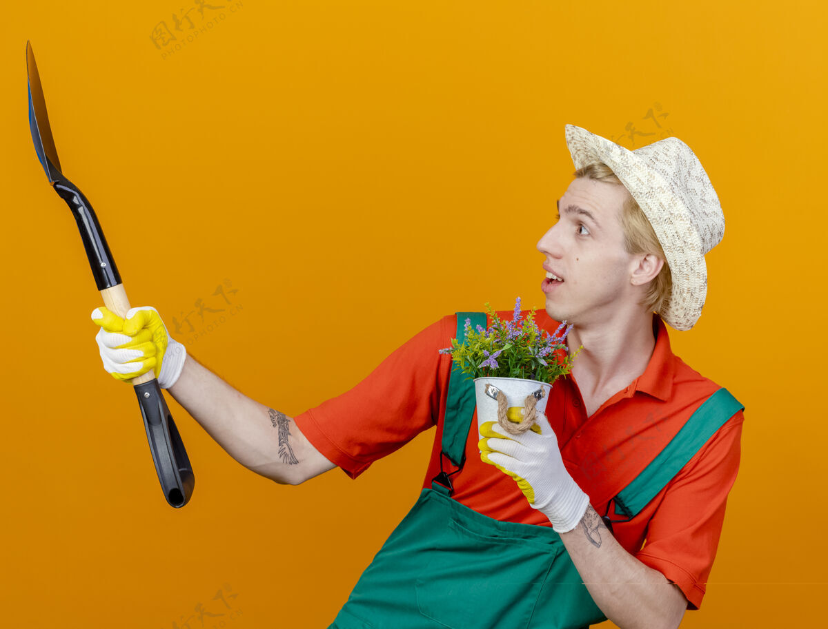 男人年轻的园丁穿着连体衣 戴着帽子 拿着铲子和盆栽植物 站在橙色的背景下惊讶地看着铲子穿橘子连身衣