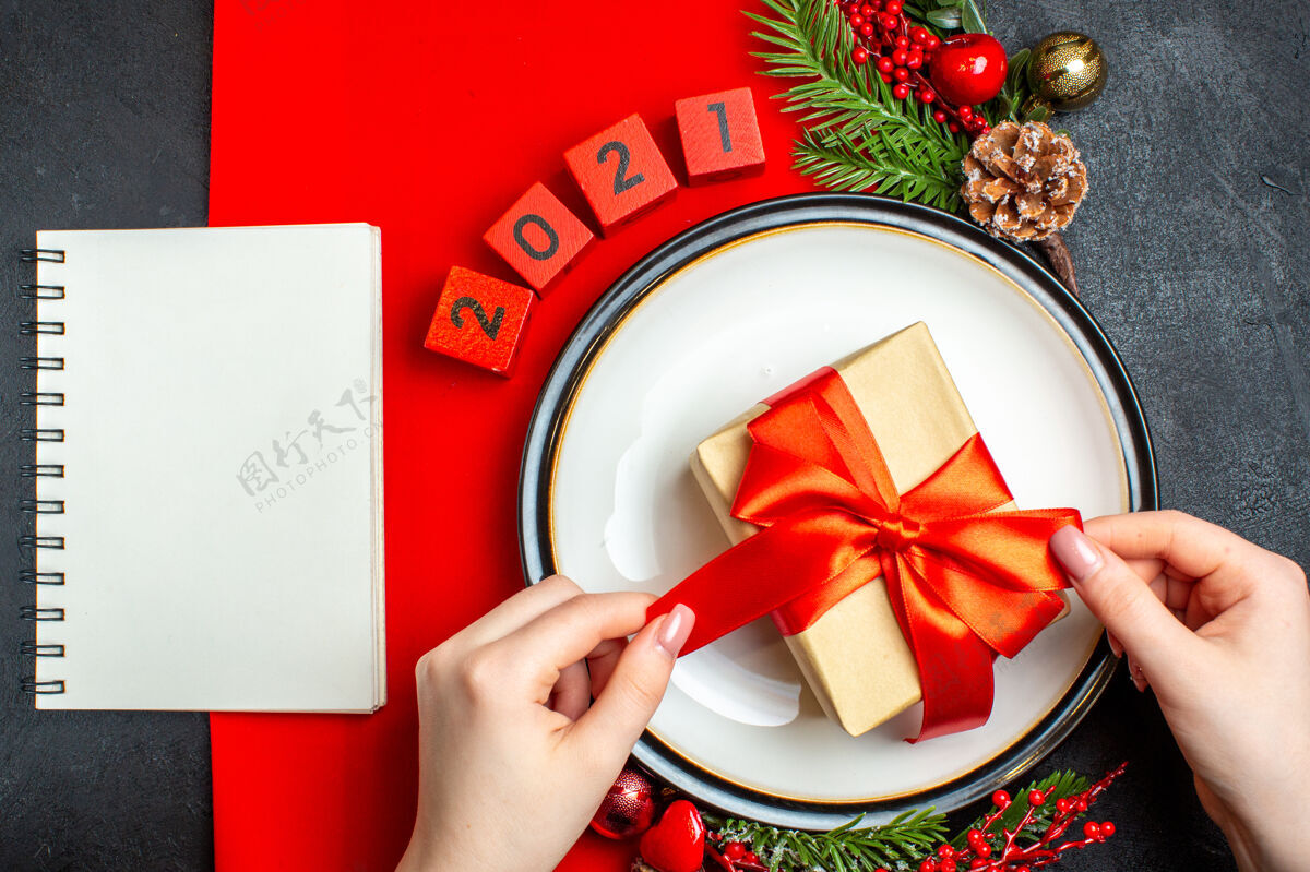 餐盘新年背景俯视图 餐盘上有礼物装饰配件 红色餐巾上有杉木树枝和数字 黑色桌子上有笔记本数字杉木头顶