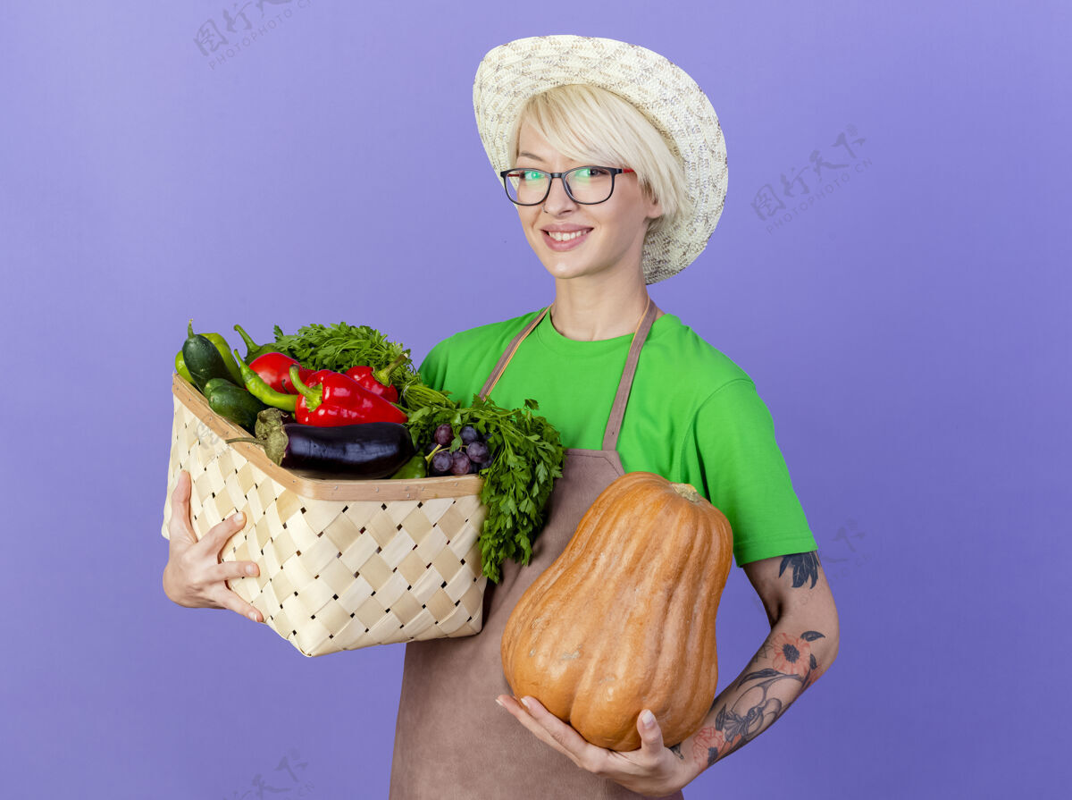 年轻围裙里留着短发 戴着帽子 拿着装满蔬菜和南瓜的箱子的年轻园丁看着镜头 站在蓝色的背景下 开心地微笑着女人短裤板条箱