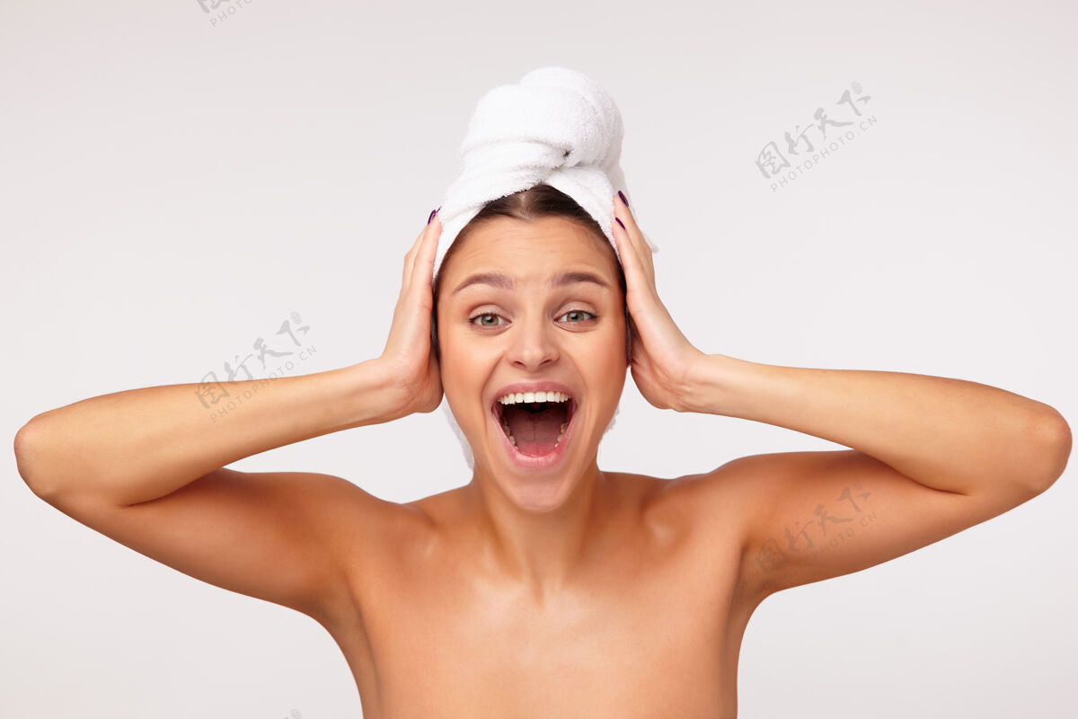 站立兴奋的年轻漂亮女士 黑发裹在浴巾里 双手紧握着头 张大嘴巴看着摄像机 与白色背景隔绝女士肩膀抬高