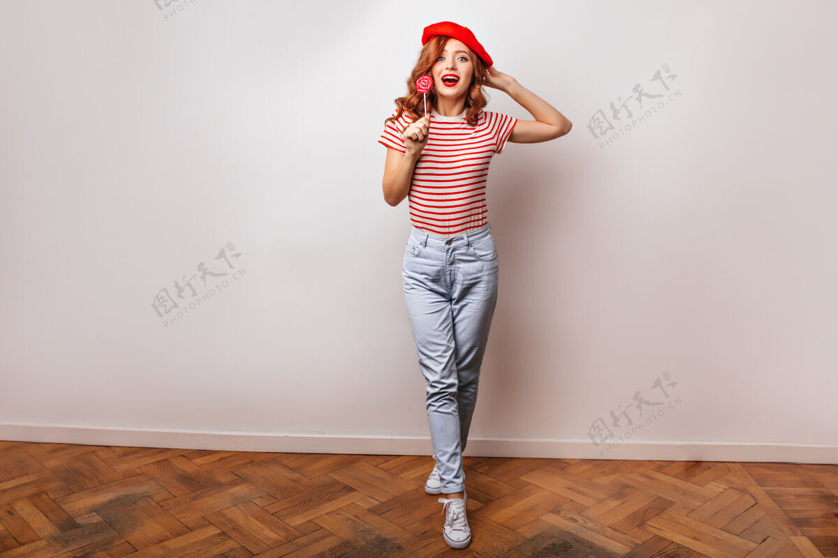姜穿着时髦牛仔裤吃棒棒糖的法国女孩的全长照片迷人的姜女拿着糖果的室内照片性感法国表情