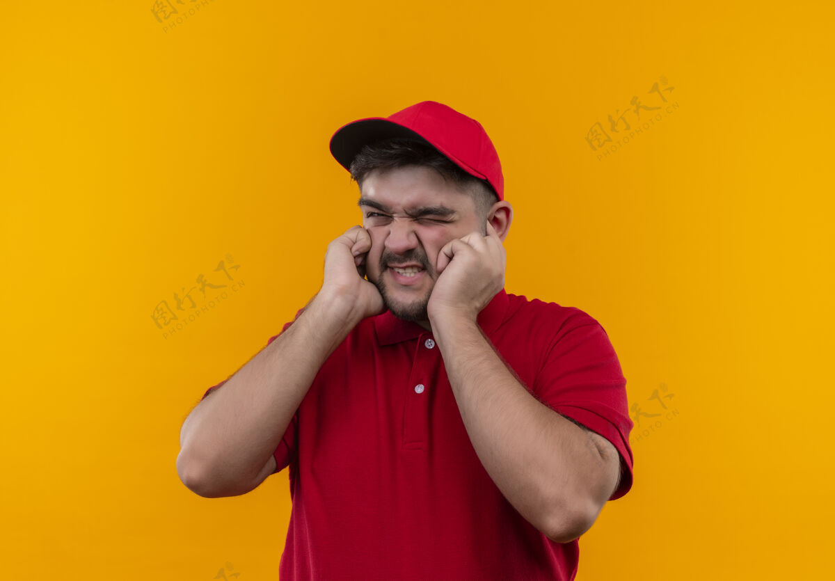 交货年轻的送货员穿着红色制服 戴着帽子 手指带着恼怒的表情看着噪音噪音恼火橙色