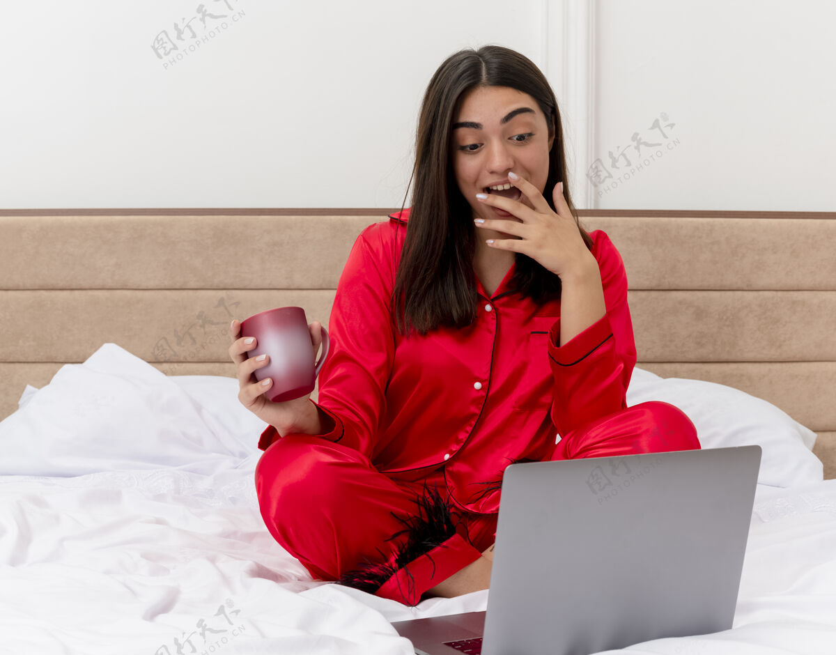 女士穿着红色睡衣的年轻美女坐在床上 手里拿着笔记本电脑和一杯咖啡 看着笔记本电脑的屏幕 在卧室内部的灯光背景下欢快地微笑着年轻微笑咖啡