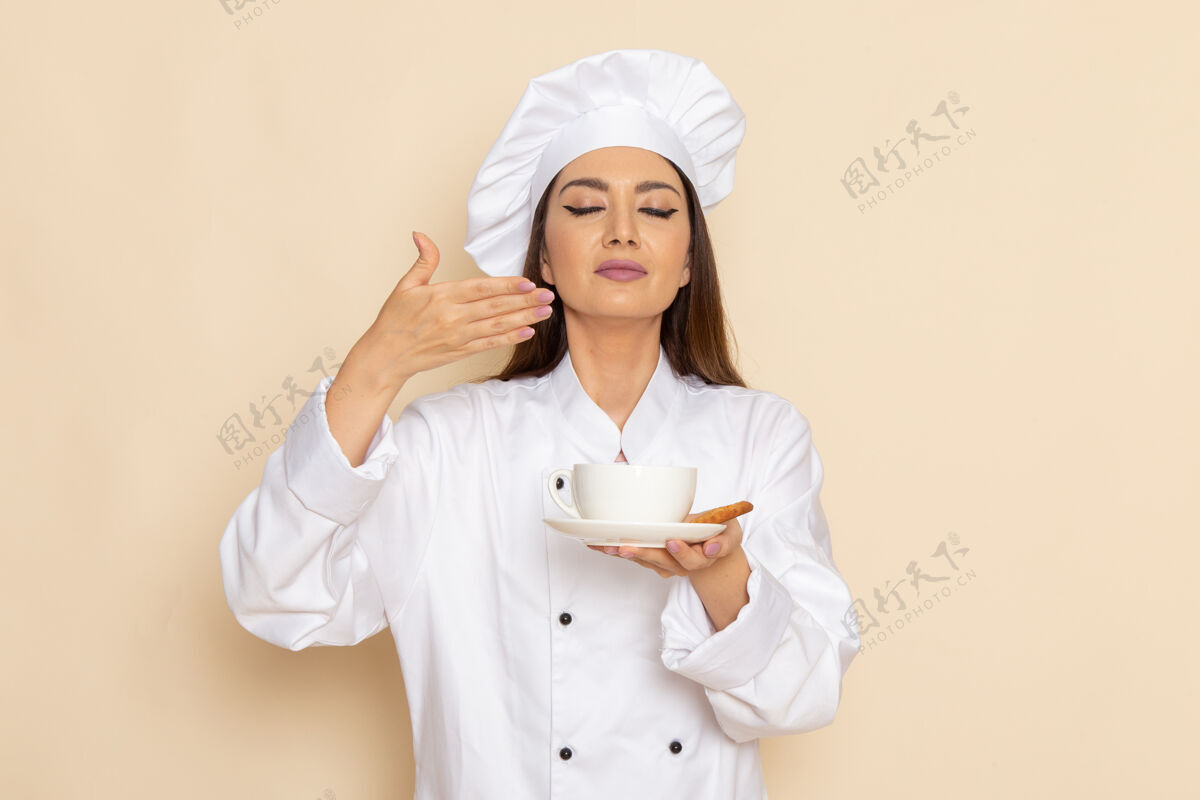 烹饪身穿白色厨师服的年轻女厨师正拿着一杯咖啡站在浅白的墙上烹饪人厨房