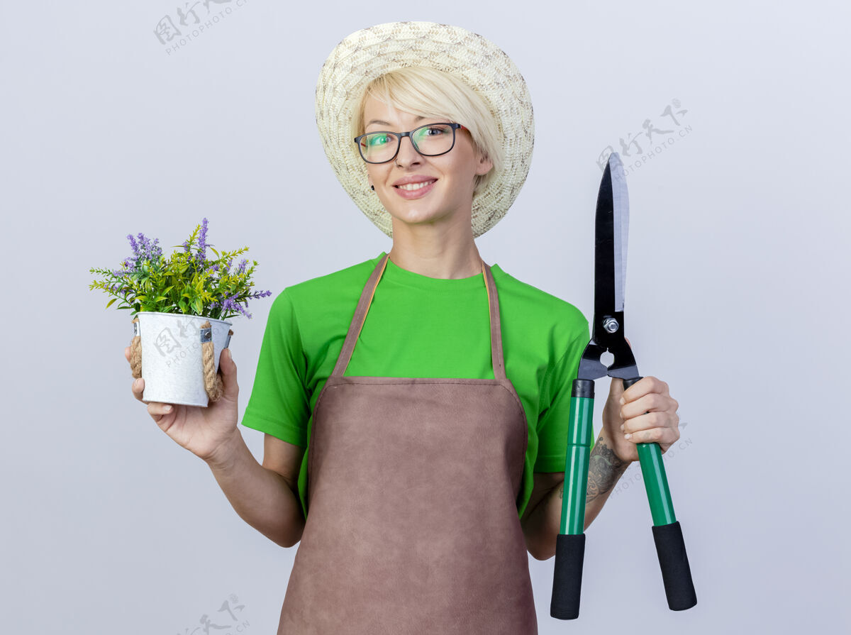 树篱一位年轻的园丁 围裙上留着短发 戴着帽子 手里拿着树篱剪和盆栽植物 站在白色的背景上 面带微笑地看着摄像机围裙帽子短裤