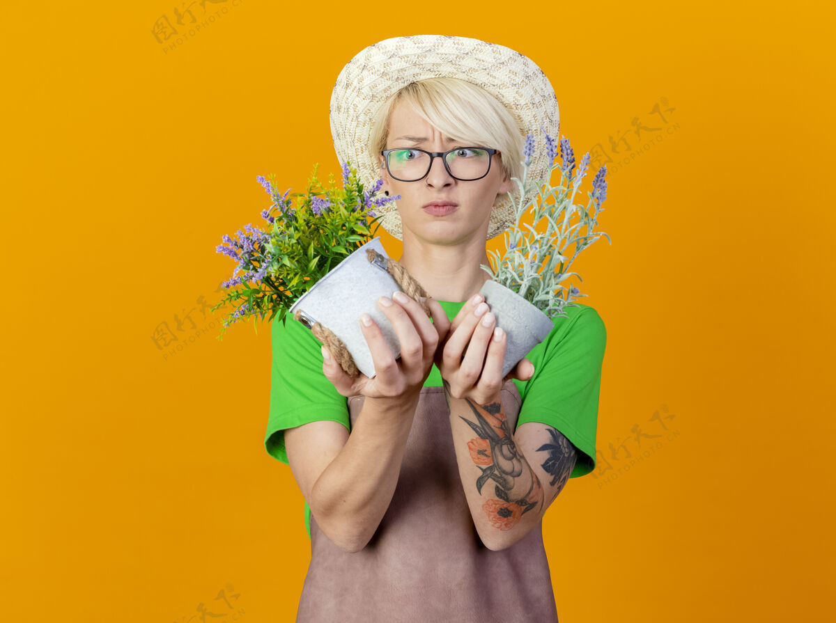 抱围裙上留着短发 戴着帽子 手里拿着盆栽植物的年轻园丁站在橙色背景下 看着它们困惑和不快围裙花园女人