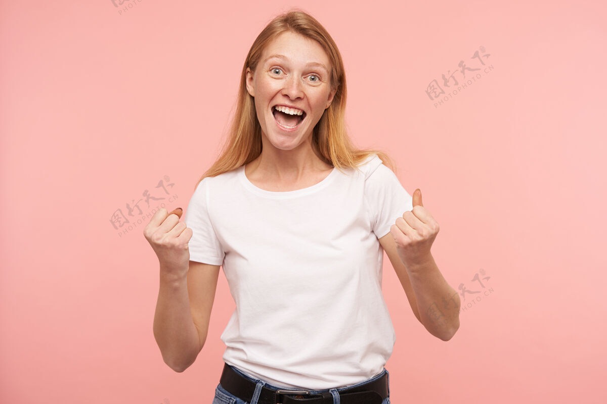 手摄影棚拍摄的年轻兴奋美丽的红发女士举起情绪化的手 同时高兴的东西 穿着基本的白色t恤 而站在粉红色的背景红发红发女人