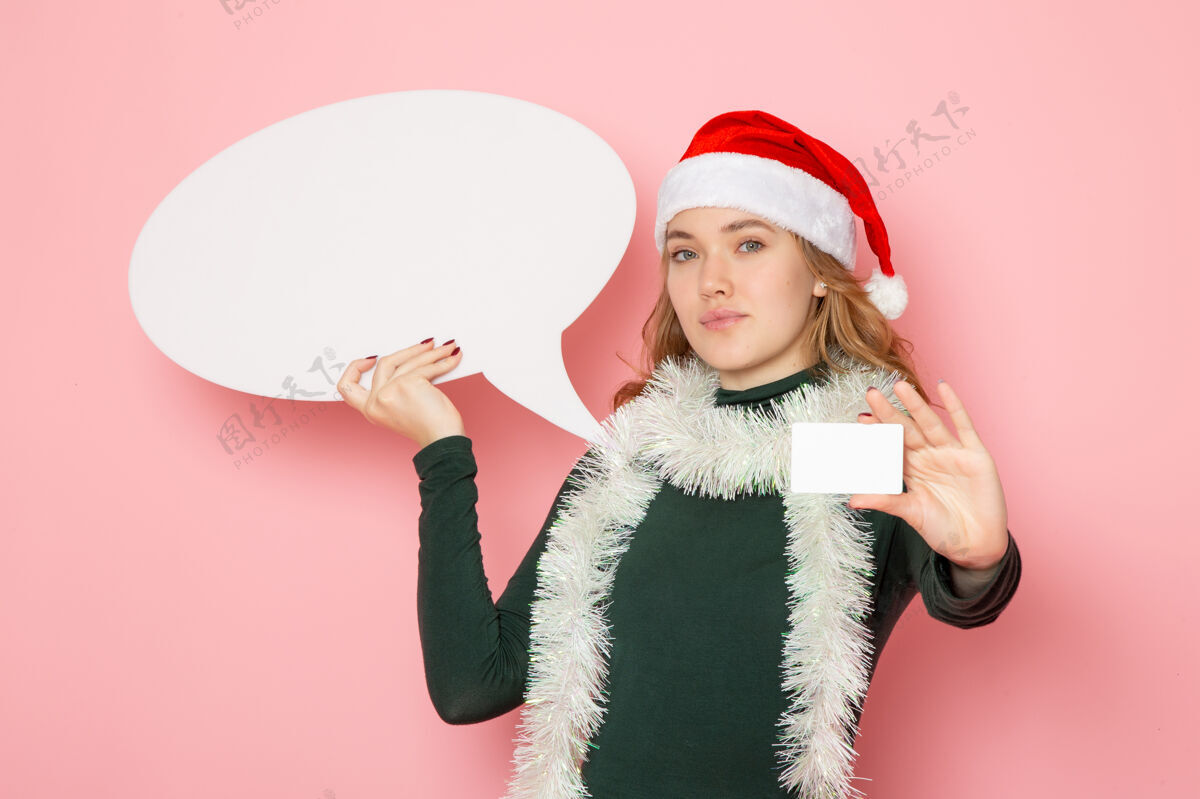 情绪正面图年轻女性手持白色大牌子和银行卡在粉红色墙上的模型圣诞节颜色帽子观点
