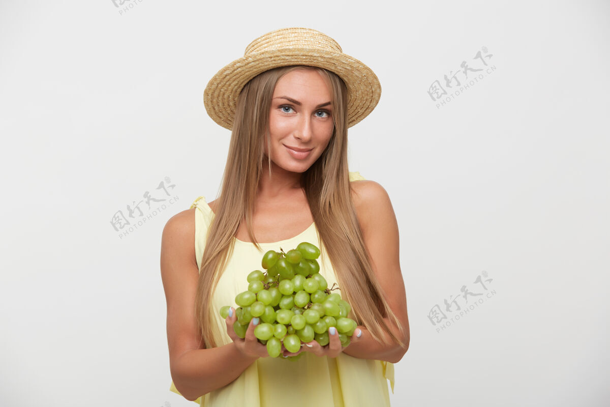 白色迷人的年轻长发金发女性 头戴船工帽 双唇合拢 正对着镜头 手持绿色葡萄 站在白色背景上束女性表情