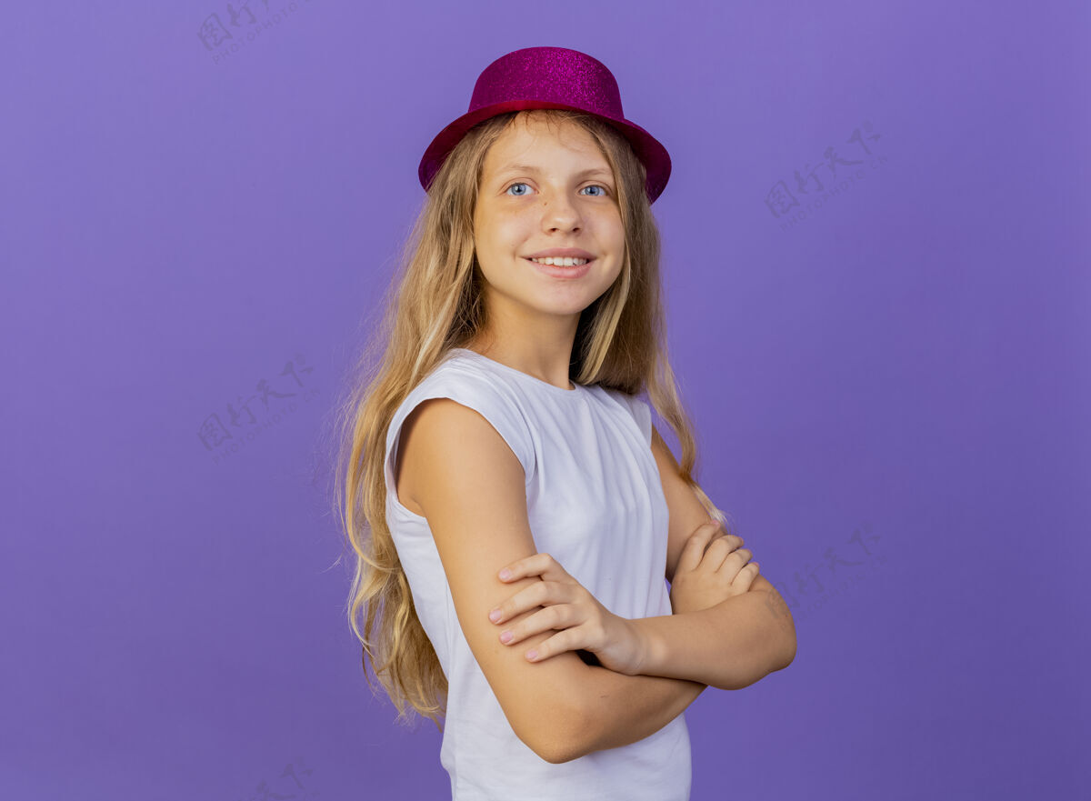 微笑戴着节日礼帽的漂亮小女孩面带微笑 双手交叉放在胸前 站在紫色背景上的生日派对概念生日十字架紫色