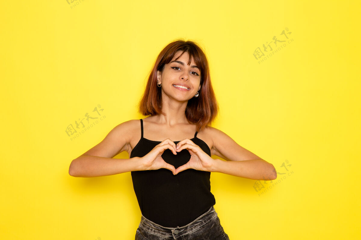 光身穿黑色衬衫的年轻女性在浅黄色墙上微笑并显示心形标志的正面视图姿势模型显示