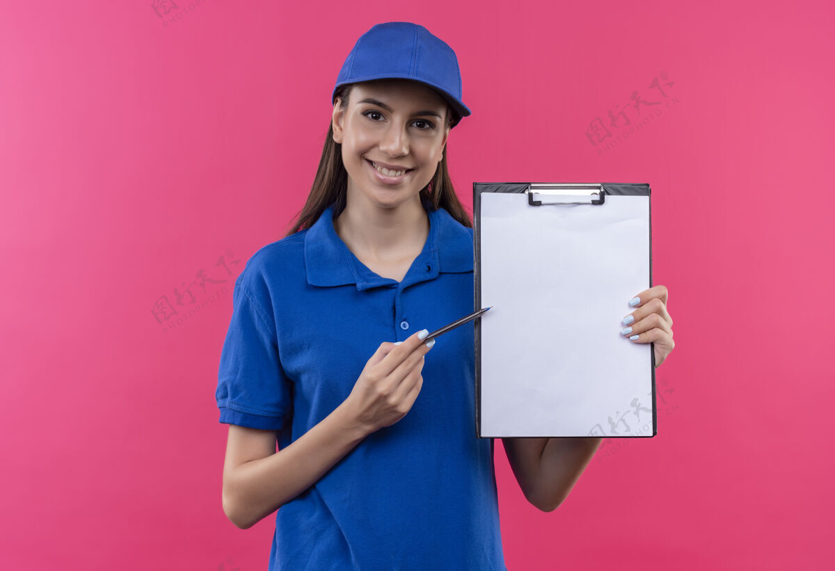 递送穿着蓝色制服 戴着帽子的年轻送货女孩拿着空白页的剪贴板 用钢笔指着剪贴板 面带微笑地要求签名钢笔站着拿着