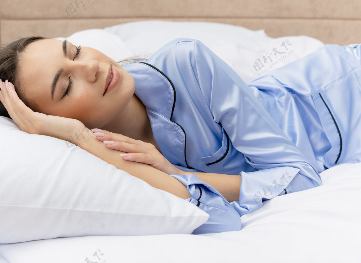 室内穿着蓝色睡衣的年轻漂亮女人躺在床上 躺在柔软的枕头上 安详地睡在家里卧室的室内灯光背景下睡眠睡衣蓝色