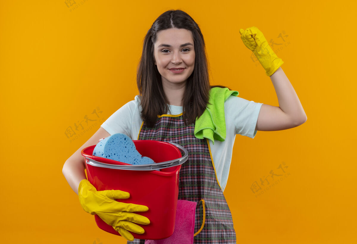 工具穿着围裙 戴着橡胶手套 拿着清洁工具的水桶的年轻漂亮女孩举起拳头 像一个微笑的胜利者微笑清洁举行