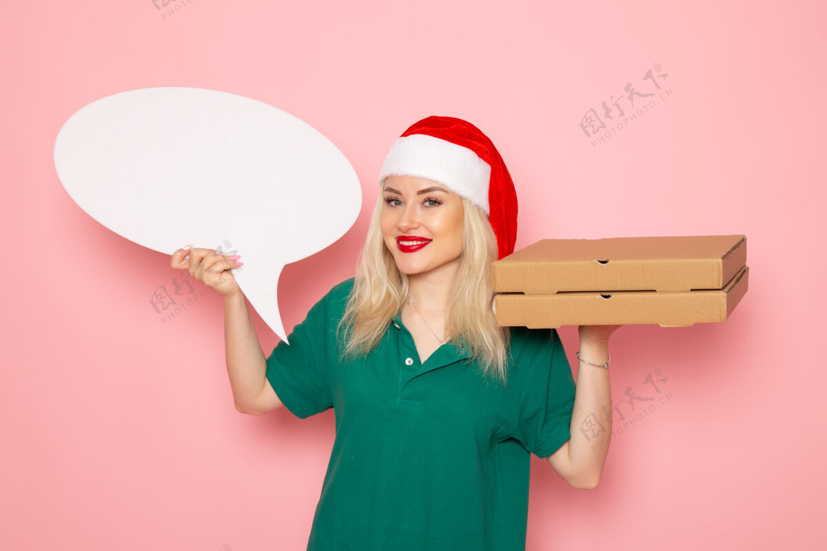 风景正面图年轻女性手持白色大牌子和食品盒在粉色墙上拍照工作制服新年假期工作快递制服大盒子