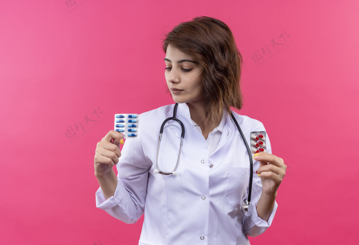 看年轻的女医生 穿着白大褂 手持听诊器 手里拿着水泡 手里拿着药丸 一脸严肃地看着它医生听诊器严重