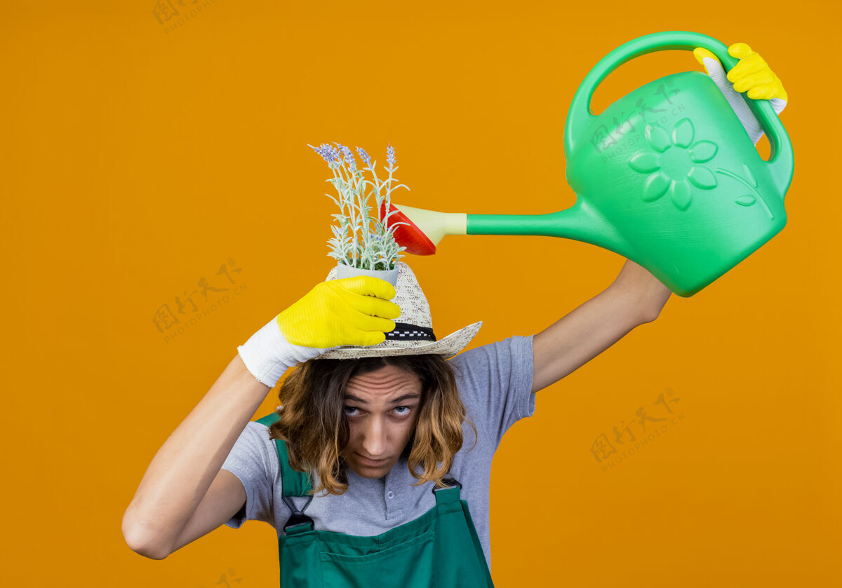 连身衣年轻的园丁戴着橡胶手套 穿着连体衣 戴着帽子 手里拿着浇水罐 站在橙色的背景上浇灌着他头上的植物橘子站着帽子