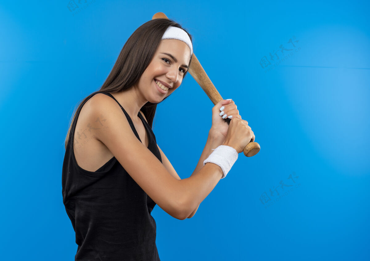运动微笑的年轻漂亮的运动女孩戴着头带和手环拿着棒球棒孤立在蓝色的空间蓝色穿年轻