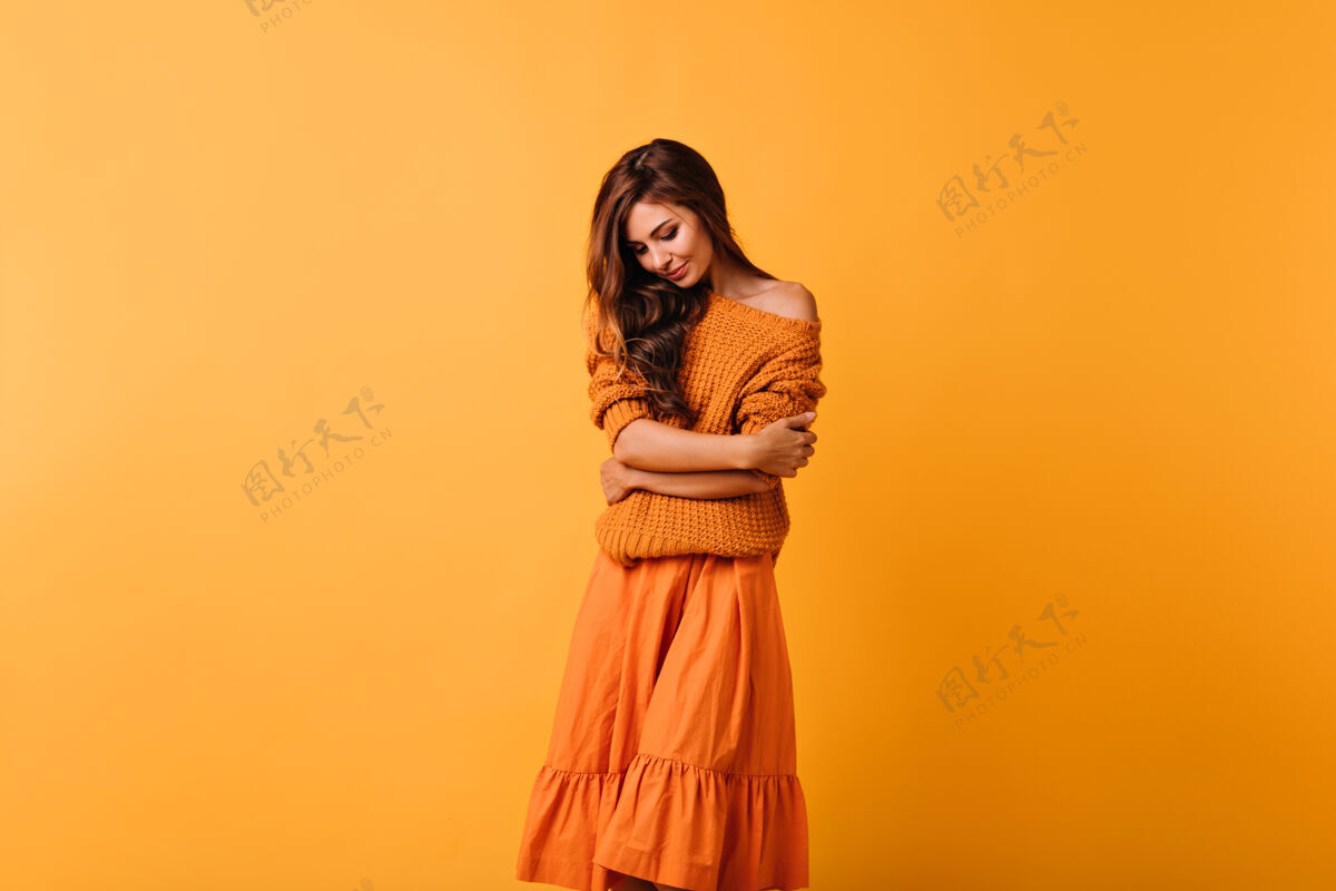服装浪漫的黑发女士穿着舒适的毛衣在黄色的床上摆姿势幽默的女模特穿着橙色的裙子往下看明亮成人时尚