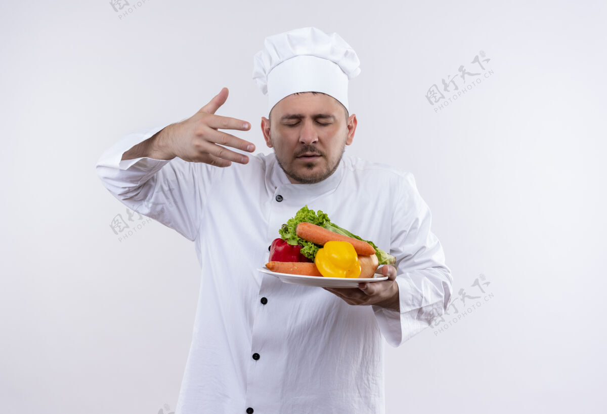 眼睛年轻帅气的厨师穿着厨师制服 拿着盛着蔬菜的盘子 举起手 闭上眼睛 在与世隔绝的白色空间里嗅着蔬菜制服封闭烹饪