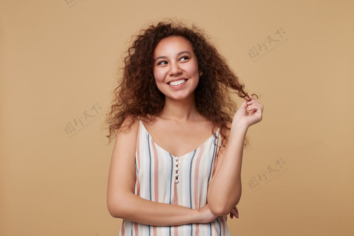 姿势快乐的年轻漂亮的卷发黑发女士的肖像 身着夏季束带上衣 一边愉快地微笑 一边梦幻般地看着一边 孤立在米色上休闲欧式女性