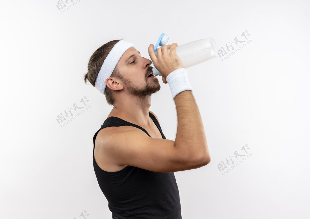 风景年轻英俊的运动型男子戴着头带和腕带站在侧视图和饮用水从瓶子隔离在白色空间年轻瓶子头带