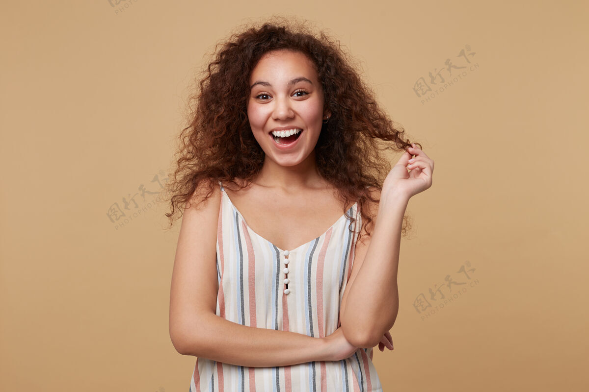 女士快乐的年轻可爱的棕色头发卷曲的女性一边玩着她的头发 一边带着深情的微笑看着 站在米色的地板上抬高卷发条纹