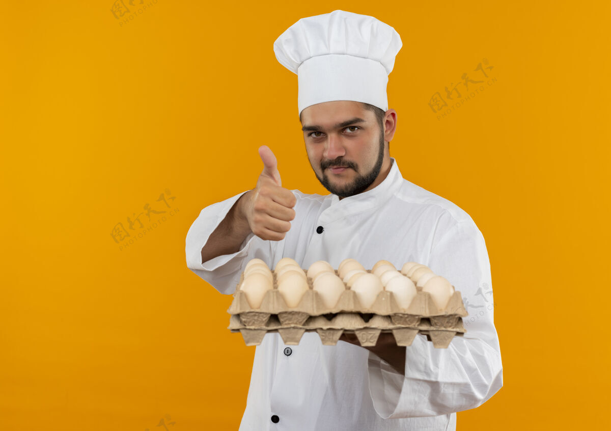 厨师身着厨师制服的年轻男厨师手捧一盒鸡蛋 在橙色的空间里孤立地竖起大拇指年轻空间男性
