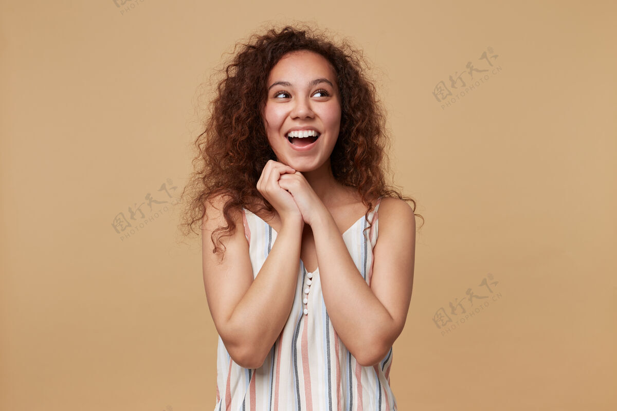 牙齿快乐的年轻可爱的棕色头发卷曲的女性 双手叠在脸下 开心地看着一边 面带微笑 站在米色的椅子上自然女人快乐