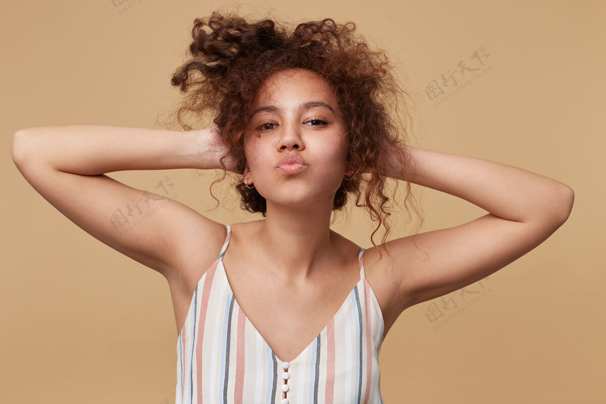女性正面的年轻棕色头发卷曲的女性肖像 双手举在头上 噘着嘴唇 孤立在米色上自然女人条纹