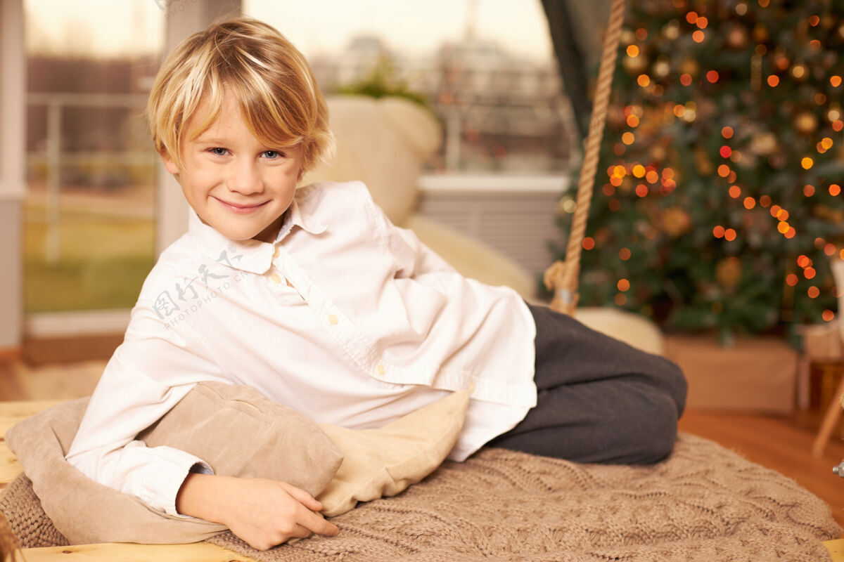 孩子室内拍摄的十岁帅气可爱的男孩 整齐的发型和快乐的笑容摆在枕头上 躺在装饰着玩具和花环的圣诞树前的地板上童年和节日男孩夏娃肖像
