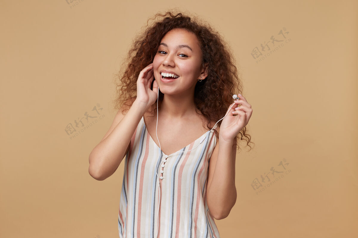 化妆年轻可爱的长发卷曲的黑发女子的肖像 戴着耳机听音乐时开心地微笑着 孤立在米色上米色衣服站立