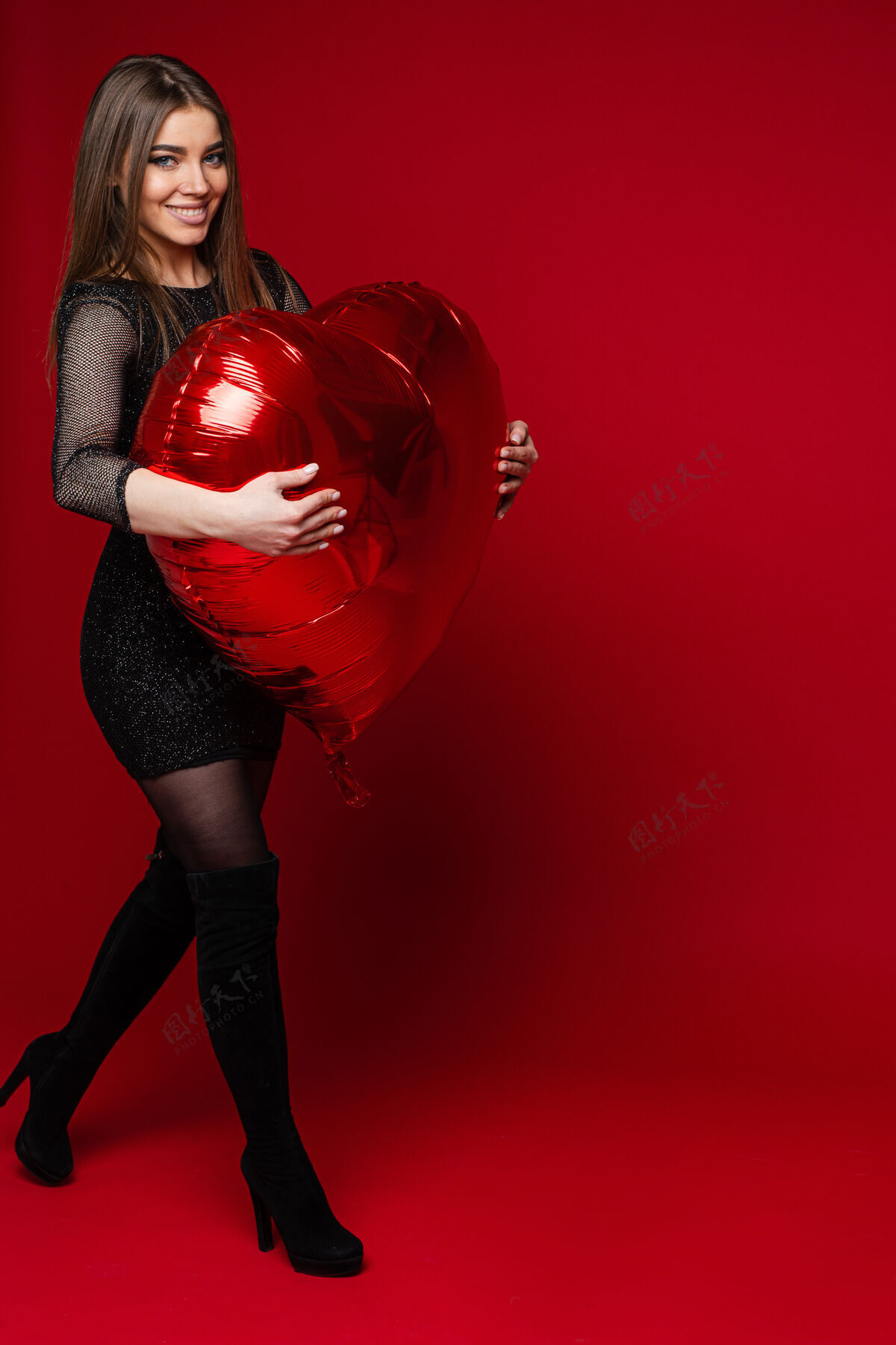 女人穿着黑色连衣裙和高筒靴 穿着红色充气心形大气球的美丽快乐的黑发女郎的全长肖像庆祝天2月14日