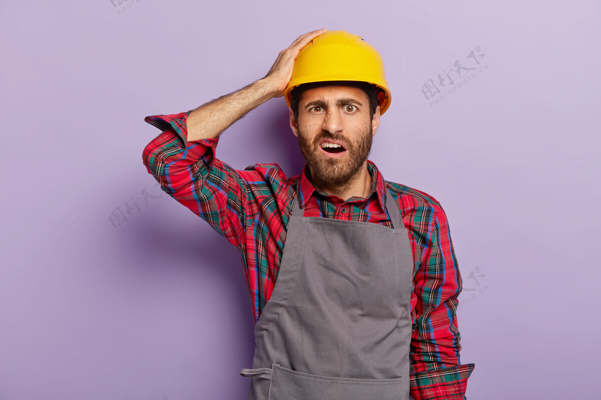 工头维修 施工和维护理念不满的未刮胡子的杂工戴着黄色防护帽 围裙 衬衫 干体力活施工人员表情消极不高兴头饰站立
