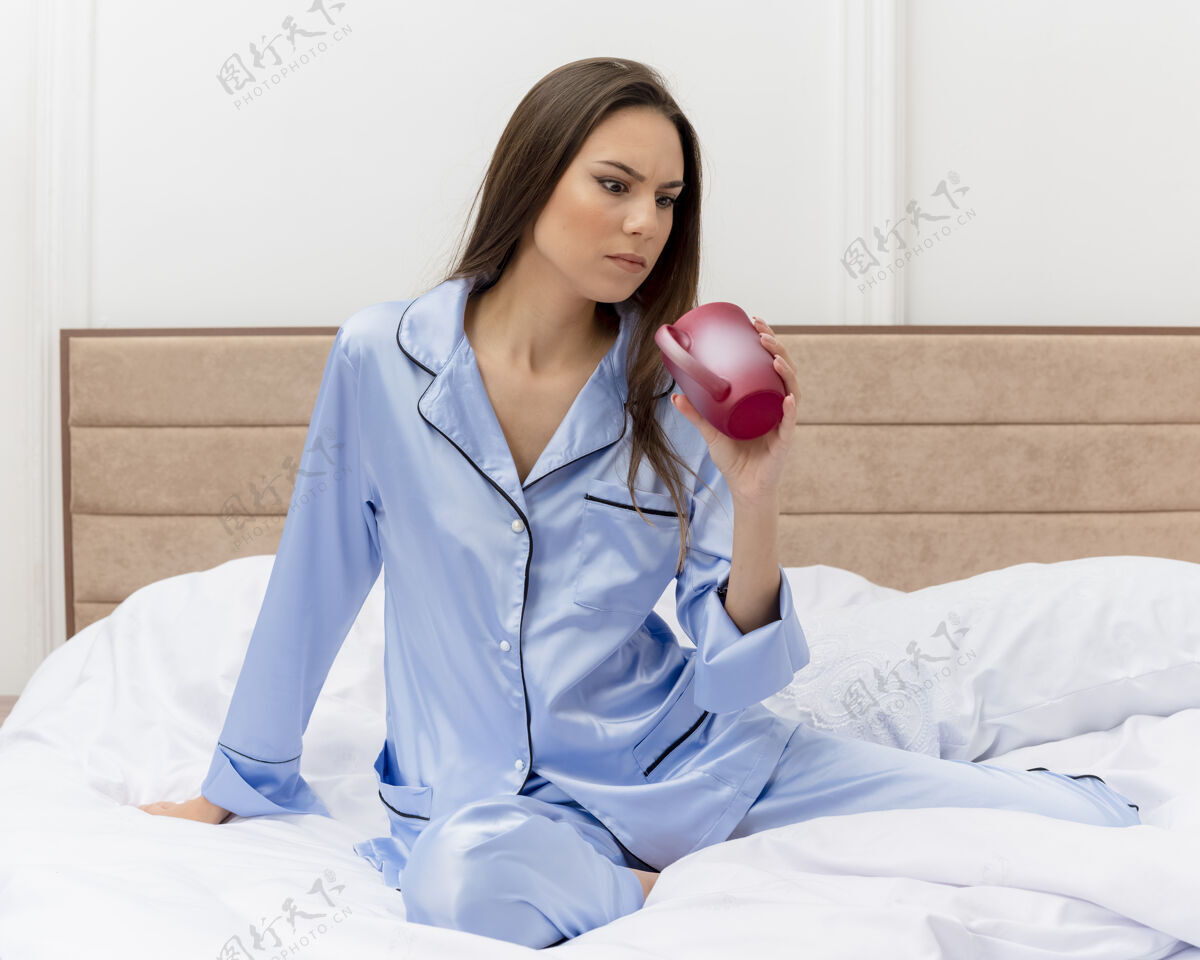 女士穿着蓝色睡衣的年轻美女坐在床上 端着一杯咖啡 看着茶杯不高兴地躺在卧室的室内灯光背景下杯子不高兴年轻