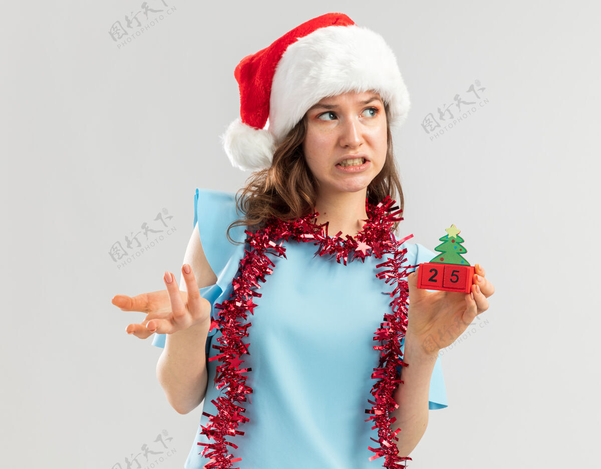 立方体身穿蓝色上衣 戴着圣诞帽 脖子上戴着金属丝的年轻女子 手里拿着玩具立方体 手里拿着圣诞节的日期 一边困惑地看着一边 一边不高兴地伸出手臂帽子旁边年
